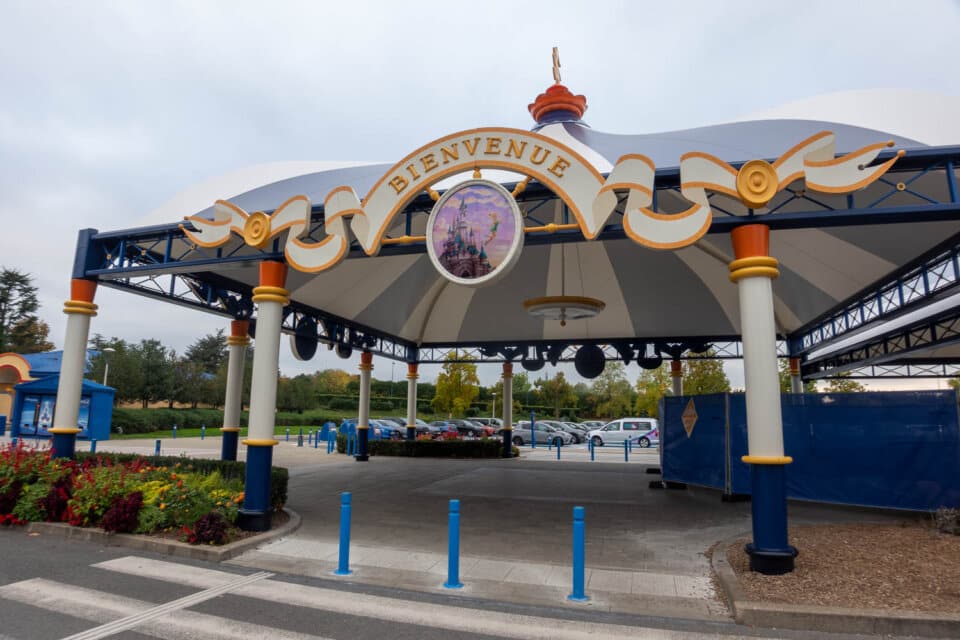 Eingangsbereich vom Parkplatz des Disneyland Paris mit überdachter Struktur, einem ‚Bienvenue‘-Schild und einem Emblem mit dem Schloss.